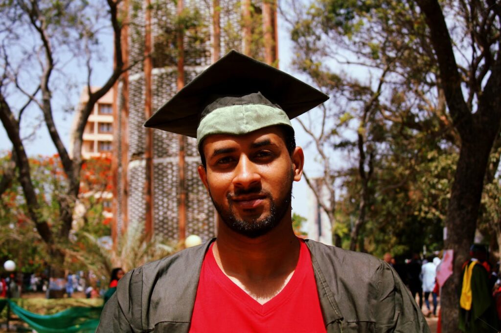 A man in a graduation cap and gown smiles confidently in an outdoor setting.